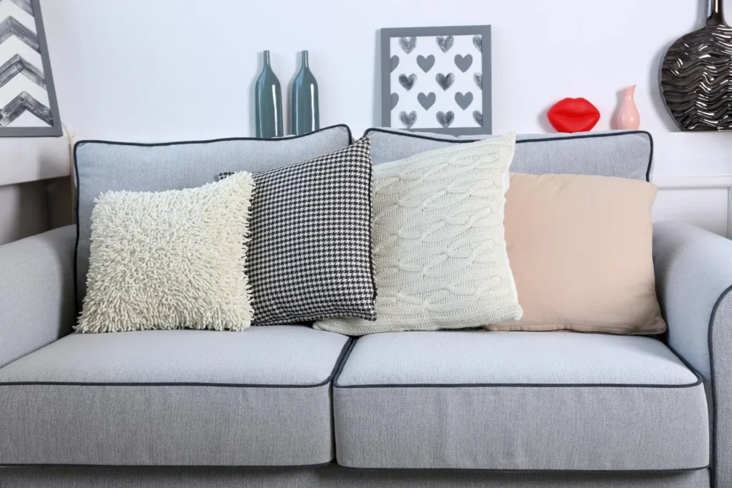 Combinación de cojines con distintas texturas (borreguito, tejido de cuadros, punto trenzado y algodón liso) sobre un sofá gris, ejemplo de cómo aportar profundidad y estilo
